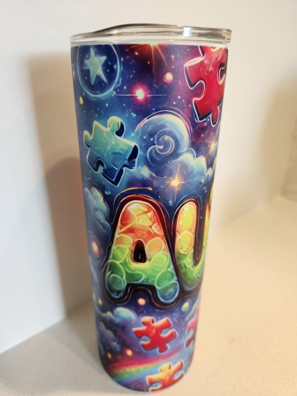 Autistic Pride glow in the dark tumbler - front view showing AU in bold 3D rainbow letters with puzzle pieces on galaxy background